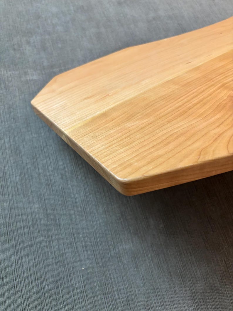 May include: Close-up of a light-colored wooden cutting board with a unique, angled shape. The wood grain is visible, and the edges are rounded. The board is likely used for food preparation and serving.