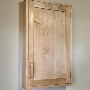 Pode incluir: Um armário de madeira clara com uma porta retangular e uma maçaneta. O armário é montado na parede e tem grão de madeira visível. A porta é emoldurada com uma borda de madeira mais escura.
