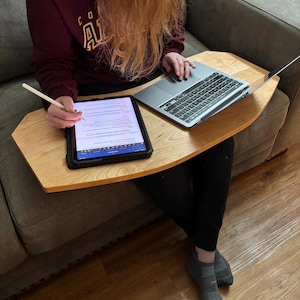 May include: A light-colored wooden lap desk with a tablet and laptop on top. The tablet is black with a white screen, and the laptop is silver. A stylus is held near the tablet. The desk is on a gray sofa.