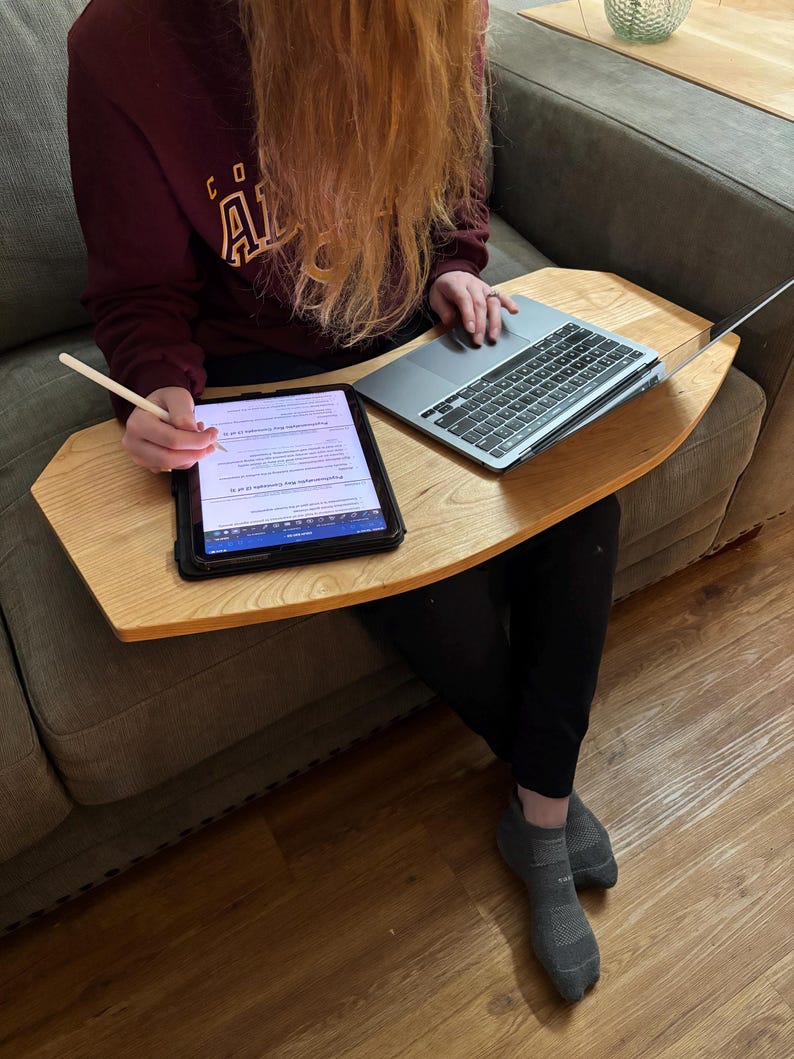 May include: A light-colored wooden lap desk with a tablet and laptop on top. The tablet is black with a white screen, and the laptop is silver. A stylus is held near the tablet. The desk is on a gray sofa.