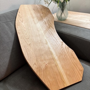 May include: A light-colored wooden serving tray with a unique, curved design. The tray is made of natural wood, showcasing the grain and texture. It rests on a gray sofa, suggesting its use for serving or display. The tray's shape is ideal for placing on a sofa arm.