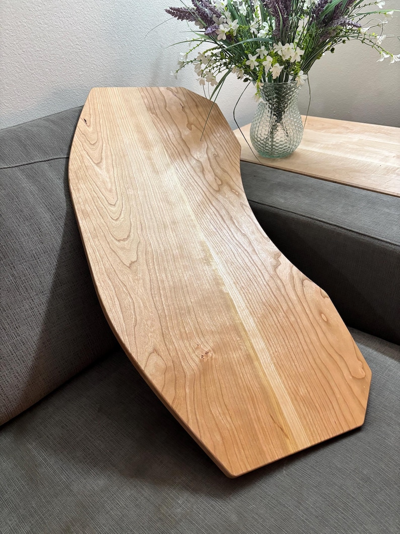 May include: A light-colored wooden serving tray with a unique, curved design. The tray is made of natural wood, showcasing the grain and texture. It rests on a gray sofa, suggesting its use for serving or display. The tray's shape is ideal for placing on a sofa arm.