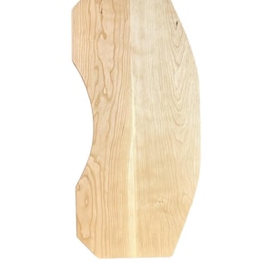 May include: A light-colored, wooden cutting board with a unique, curved design. The board has a natural wood grain pattern and a smooth surface. The shape is asymmetrical, with one side featuring a curved edge and the other side having a straight edge.