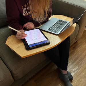 May include: A light-colored wooden lap desk with a laptop and tablet. The laptop is open, and the tablet is being used with a stylus. The desk is resting on a person's lap while they are sitting on a gray couch.