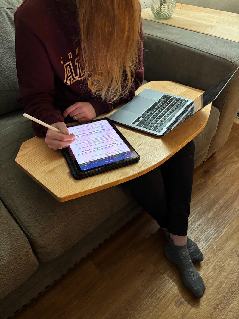 May include: A light-colored wooden lap desk with a laptop and tablet. The laptop is open, and the tablet is being used with a stylus. The desk is resting on a person's lap while they are sitting on a gray couch.