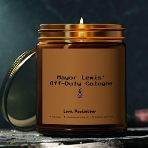 May include: A brown glass candle jar with a gold lid. The candle is lit and the label reads "Mayor Lewis' Off-Duty Cologne" and "Love, Pookiebear".