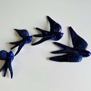May include: Four ceramic swallows in flight, each with a glossy, dark blue finish. The birds are arranged on a white surface, showcasing their detailed wing and tail designs. A decorative accent for any space.