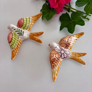 May include: Two ceramic swallow figurines with white bodies, orange wings, and decorative floral patterns. One has green and red accents, the other has brown floral details. The birds are set against a white background with pink flowers and green leaves.