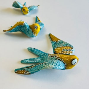 Puede incluir: Tres pájaros de cerámica en vuelo, pintados en tonos de azul y amarillo. Los pájaros están dispuestos en fila, con el pájaro más grande en primer plano y el pájaro más pequeño en el fondo.