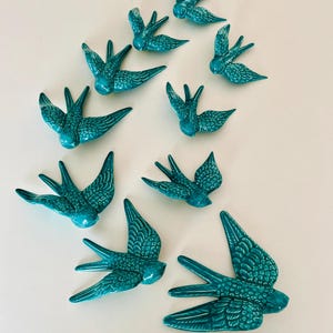 Puede incluir: Un conjunto de diez decoraciones de pared de golondrinas de cerámica azul verdoso. Las aves están dispuestas en un patrón escalonado, con el ave más grande en la parte inferior y el ave más pequeña en la parte superior.