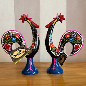 May include: Two black rooster figurines with colorful painted details. The roosters have pink combs and wattles, and their bodies are decorated with flowers and hearts. The roosters are standing on blue bases.