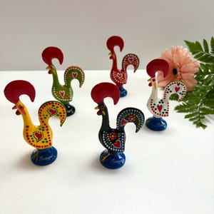 May include: A selection of hand-painted ceramic rooster figurines. Each rooster features a red comb and a blue base with the word "Portugal". The roosters are decorated with various patterns and colours, including yellow, green, black, and white.