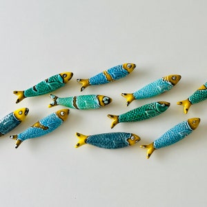 May include: Nine ceramic fish figurines in various shades of blue and green with yellow accents. The fish are arranged in a staggered pattern on a white surface.