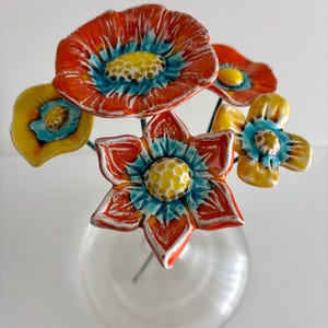 Pode incluir: Um conjunto de cinco flores de cerâmica em hastes metálicas finas, dispostas num vaso de vidro transparente. As flores apresentam esmaltes vibrantes em laranja, amarelo e turquesa com centros texturizados e pétalas com bordas brancas.