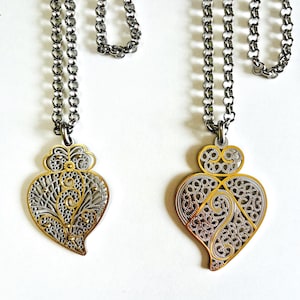 May include: Two filigree heart-shaped pendants with gold and silver tones hang from silver-toned chains. The pendants have intricate openwork designs. The background is plain white.
