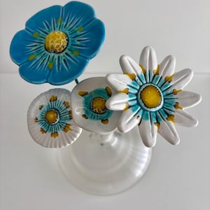 Può includere: Un set di quattro fiori in ceramica su sottili steli metallici. Un grande fiore blu con centro giallo, due fiori bianchi più piccoli con centri blu e gialli e un fiore bianco con petali lunghi, centro blu e accenti gialli.