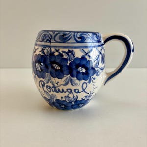 May include: A white ceramic mug with a blue floral design and the word "Portugal" in cursive. The mug has a rounded shape with a handle and is decorated with blue flowers and leaves. The rim and handle are also trimmed with blue.