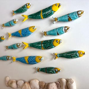 Può includere: Una collezione di sculture di pesci in ceramica di varie dimensioni e colori. I pesci presentano tonalità gialle, blu e verdi con motivi dettagliati. La disposizione suggerisce un banco di pesci che nuotano insieme, con una fila di piccole pietre in basso.