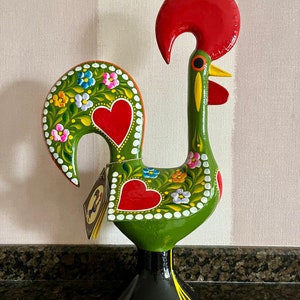 May include: A green ceramic rooster figurine featuring a large red comb, a yellow beak, and a black base. The body is decorated with hand-painted red hearts, white dots, and colorful floral patterns in blue, pink, and orange. A small paper tag hangs from the side.