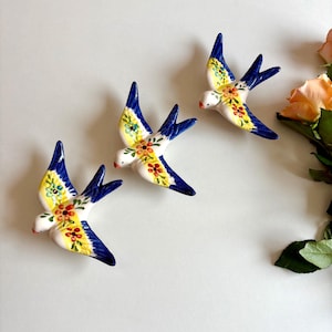Könnte beinhalten: Drei Keramik-Schwalbenfiguren. Jeder Vogel hat einen weißen Körper, gelbe und floral gemusterte Flügel und blaue Flügelspitzen. Die Schwalben sind auf einer weißen Oberfläche angeordnet, mit pfirsichfarbenen Rosen im Hintergrund.