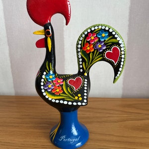 May include: A black rooster figurine with a red comb and a blue base. The rooster is painted with colourful flowers and hearts. The base is inscribed with the word "Portugal".