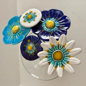 Könnte beinhalten: Eine Sammlung von fünf blumenförmigen Dekorationsobjekten aus Keramik. Die Blüten haben eine glänzende Glasur in Weiß, leuchtendem Blau und tiefem Marineblau mit strukturierten gelben Mitten. Jedes Stück ist auf einem dünnen Metallstab befestigt.