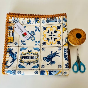 May include: A white tablecloth with a blue and yellow Portuguese tile pattern. The tablecloth has a crocheted border in a mustard yellow color. The text "Handmade in Portugal" is visible on a white tag.