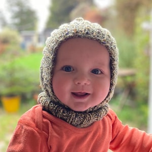 May include: A baby wearing a green and brown knitted hat with a hood. The hat has a cable knit pattern and is pulled down over the baby's ears. The baby is smiling and looking at the camera.