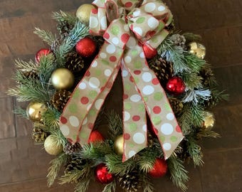 Op de afbeelding: Een feestelijke kerstkrans met een mix van gouden en rode ornamenten, dennenappels en kunstmatige dennentakken. De krans is versierd met een grote strik met een bruine achtergrond met witte, rode en groene stippen.