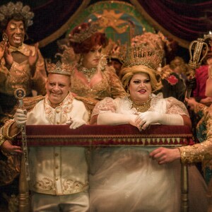 May include: A group of people dressed in elaborate costumes and crowns, including a person wearing a white gown and a crown, standing in front of a red velvet curtain.