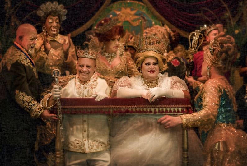 May include: A group of people dressed in elaborate costumes and crowns, including a person wearing a white gown and a crown, standing in front of a red velvet curtain.