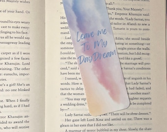 May include: A rectangular bookmark with rounded corners featuring a watercolor wash in shades of blue, purple, and peach. The text "Leave me To My Day Dream" is printed in a blue script font across the center of the bookmark surface.