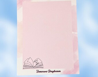May include: A rectangular notepad with a pale pink centre and a soft pink watercolour border. The bottom left features a black line drawing of an open book with the text Just one more chapter. The bottom centre displays the text Tomorrow Daydream.