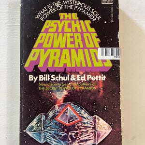 May include: A paperback book titled The Psychic Power of Pyramids by Bill Schul and Ed Pettit. The black cover features a space-themed background with stars, a glowing crystalline pyramid graphic, and bold yellow and purple text throughout.