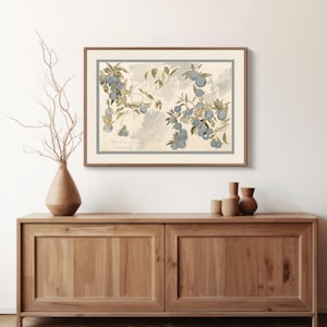 Puede incluir: Una impresión botánica enmarcada con una ilustración detallada de ciruelas en una rama, con un borde azul claro. La obra de arte se exhibe sobre un mueble de madera, junto a un jarrón decorativo y pequeñas piezas de cerámica.