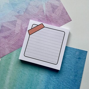 May include: A square, lined notepad with a black border and a peach-coloured tab. The notepad is on a white surface, with colourful watercolour paper in the background. The watercolour paper has shades of blue, purple, and teal.