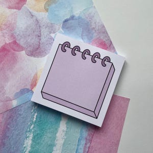 May include: A purple notepad with a spiral binding. The notepad is on a watercolor background with pink, blue, and yellow colors.