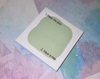 May include: A square notepad with a green blob design and the text "1. Make the plan" and "2. Have a nap" in black. The notepad is on a colorful watercolor background with shades of blue, purple, and pink.