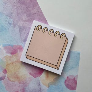 May include: A white notepad with a beige notepad illustration. The notepad illustration has a ring binding with gold rings.