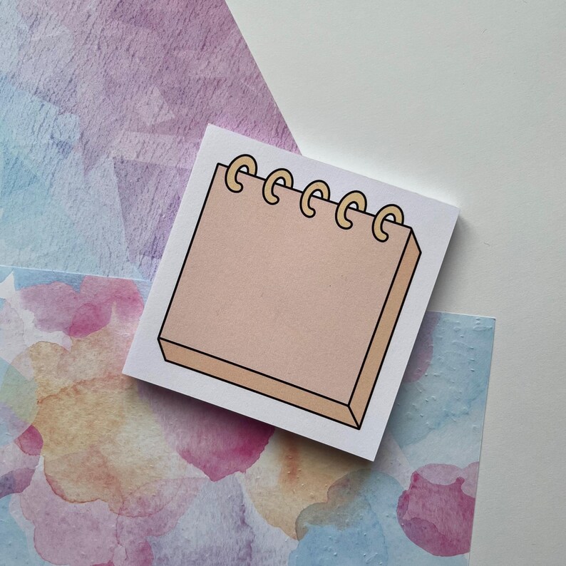 May include: A white notepad with a beige notepad illustration. The notepad illustration has a ring binding with gold rings.