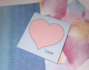 May include: A stack of light blue sticky notes with a large pink heart design and the word "Lover" printed in black. The notes are on a background of watercolor paper in shades of blue, pink, and yellow.