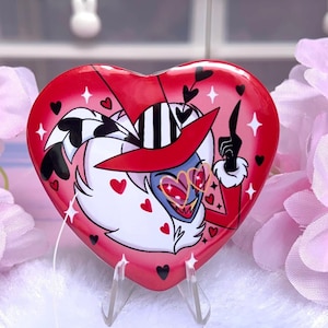 May include: A red heart-shaped button with a cartoon character wearing a red and black striped hat and sunglasses. The character has white fur and a red mouth. The background is pink with white hearts.