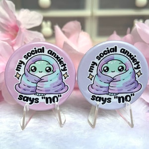 May include: Two round pink and purple buttons with a cartoon alien wrapped in a blanket. The text on the buttons reads "My social anxiety says "no""