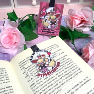 Op de afbeelding: Een roze en witte magnetische bladwijzer met een cartoonvogel die een rood-wit geruit overhemd, een kroon en een hoge hoed draagt. De bladwijzer heeft de tekst "Ask Me About My Hyperfixation" erop.