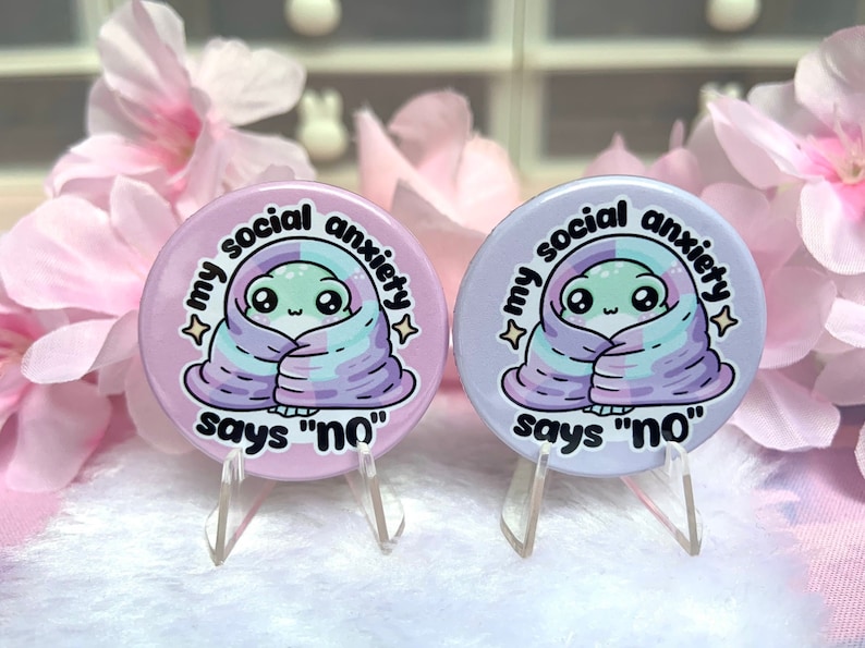 May include: Two round pink and purple buttons with a cartoon alien wrapped in a blanket. The text on the buttons reads "My social anxiety says "no""