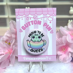 Könnte beinhalten: Eine rosa Anstecknadel mit einer Regenbogen-Teekanne und einem Frosch darin. Der Text auf der Nadel lautet "Proud to be Queer".