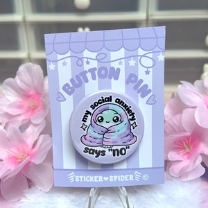 May include: A white button pin with a cartoon alien wrapped in a purple and blue blanket. The text on the pin reads "My social anxiety says "no"".