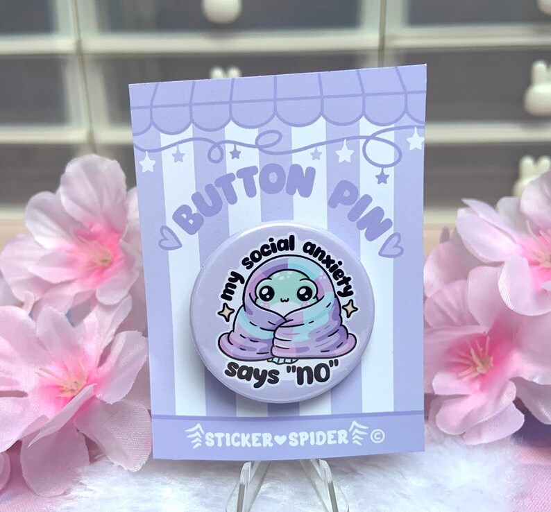 May include: A white button pin with a cartoon alien wrapped in a purple and blue blanket. The text on the pin reads "My social anxiety says "no"".