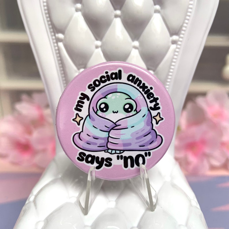 May include: A pink button with a cartoon alien wrapped in a purple and blue blanket. The text on the button reads "My social anxiety says "no"".