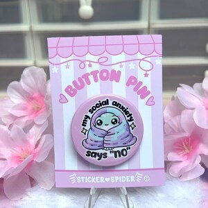 May include: A pink button pin with a cartoon alien wrapped in a purple and blue blanket. The text on the pin reads "My social anxiety says "no"".